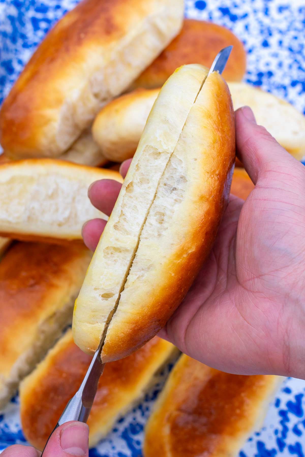 Hand holding a serrated knife slicing a sourdough hot dog bun open.