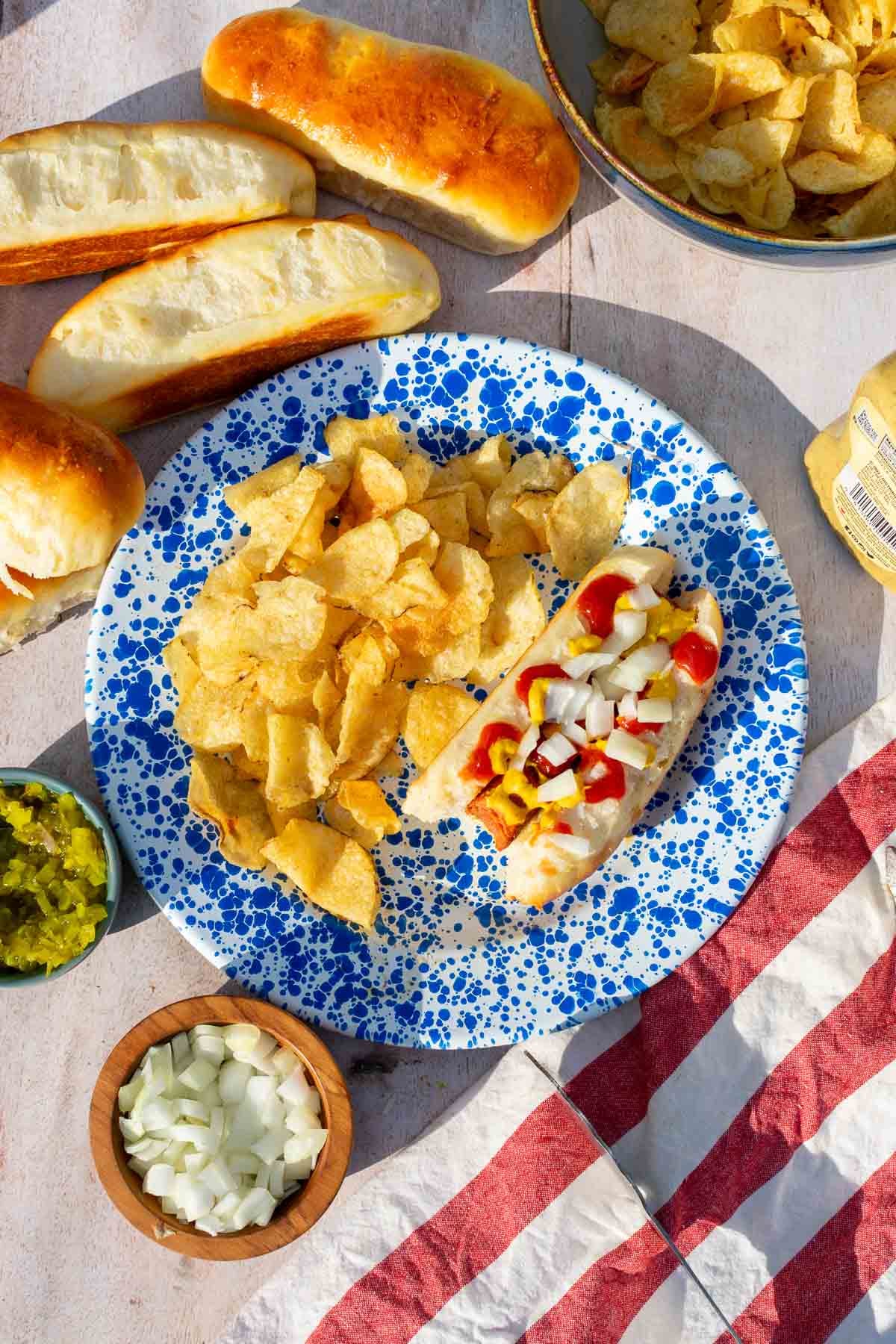 Hot dog with ketchup and mustard in a sourdough hot dog bun on a plate with potato chips and more sourdough hot dog buns and condiments around it. 