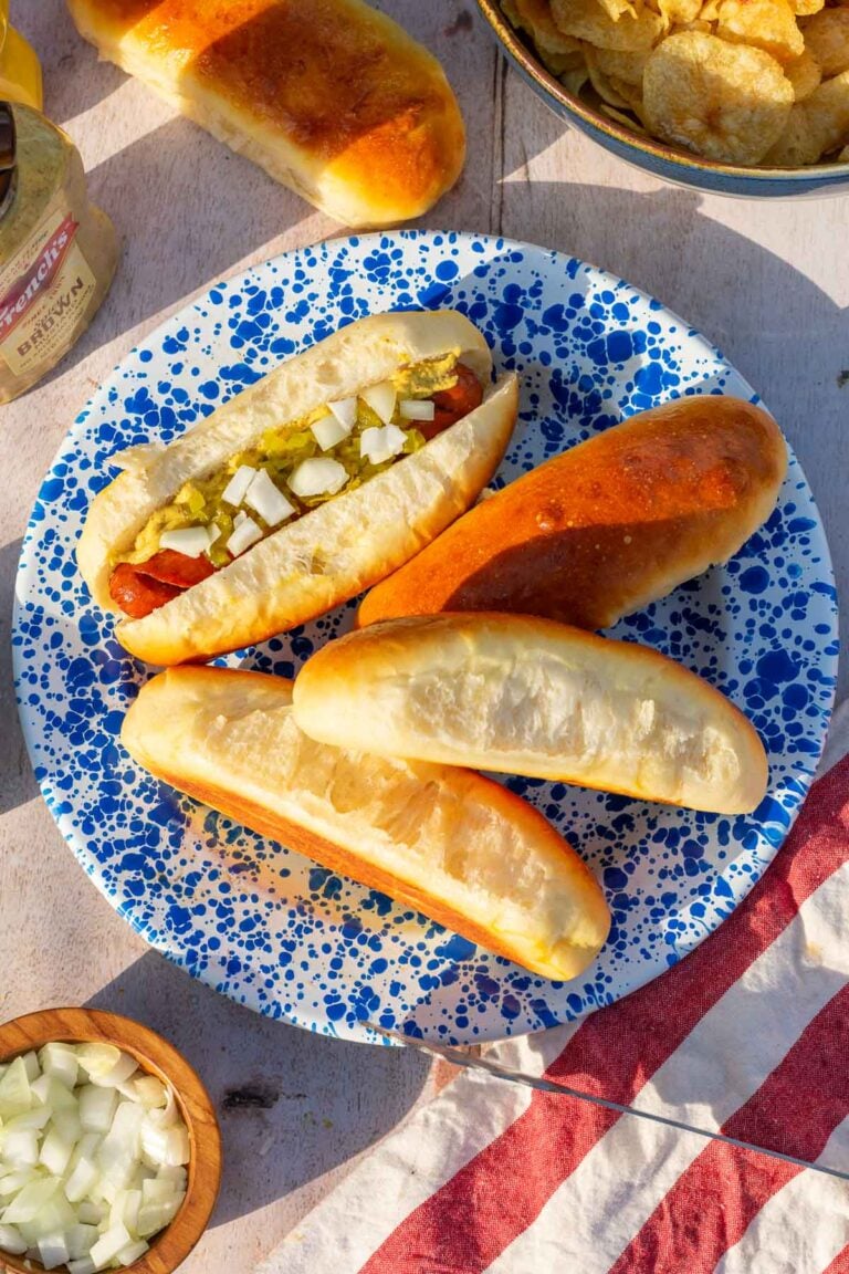 Sourdough hot dog buns on a blue splatter enamelware plate. One is filled with a hot dog with mustard, relish, and onions.