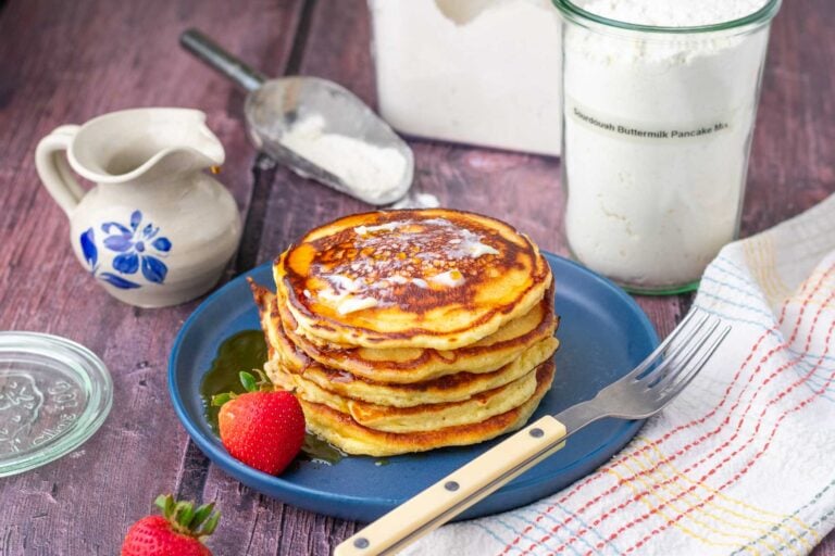 Stack of sourdough buttermilk pancakes on a plate with maple syrup with a large container of homemade sourdough pancake mix in the background with a flour scoop.