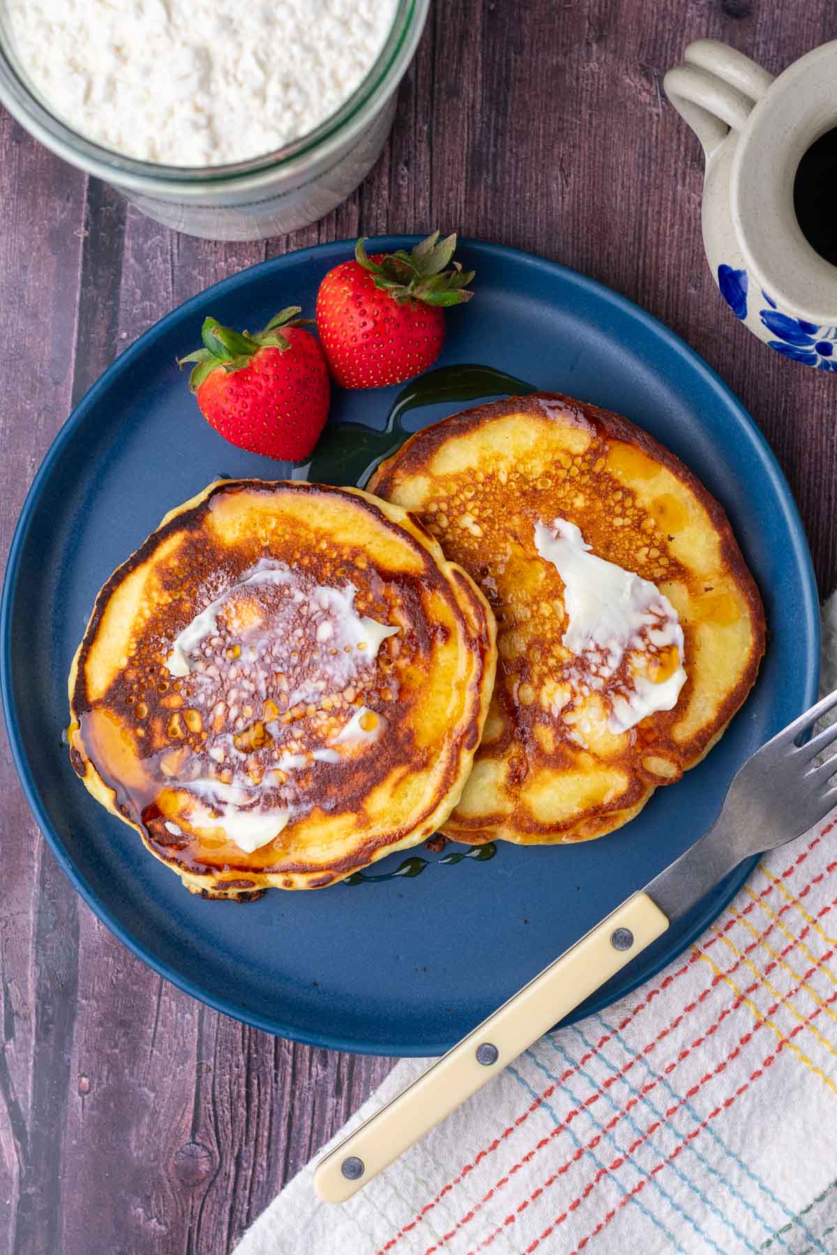 Sourdough buttermilk pancakes on a plate with butter, maple syrup, and strawberries with a fork to the side.