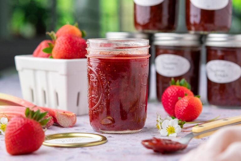 Mason jar with strawberry rhubarb jam with canned jam in the background and fresh strawberries and rhubarb.
