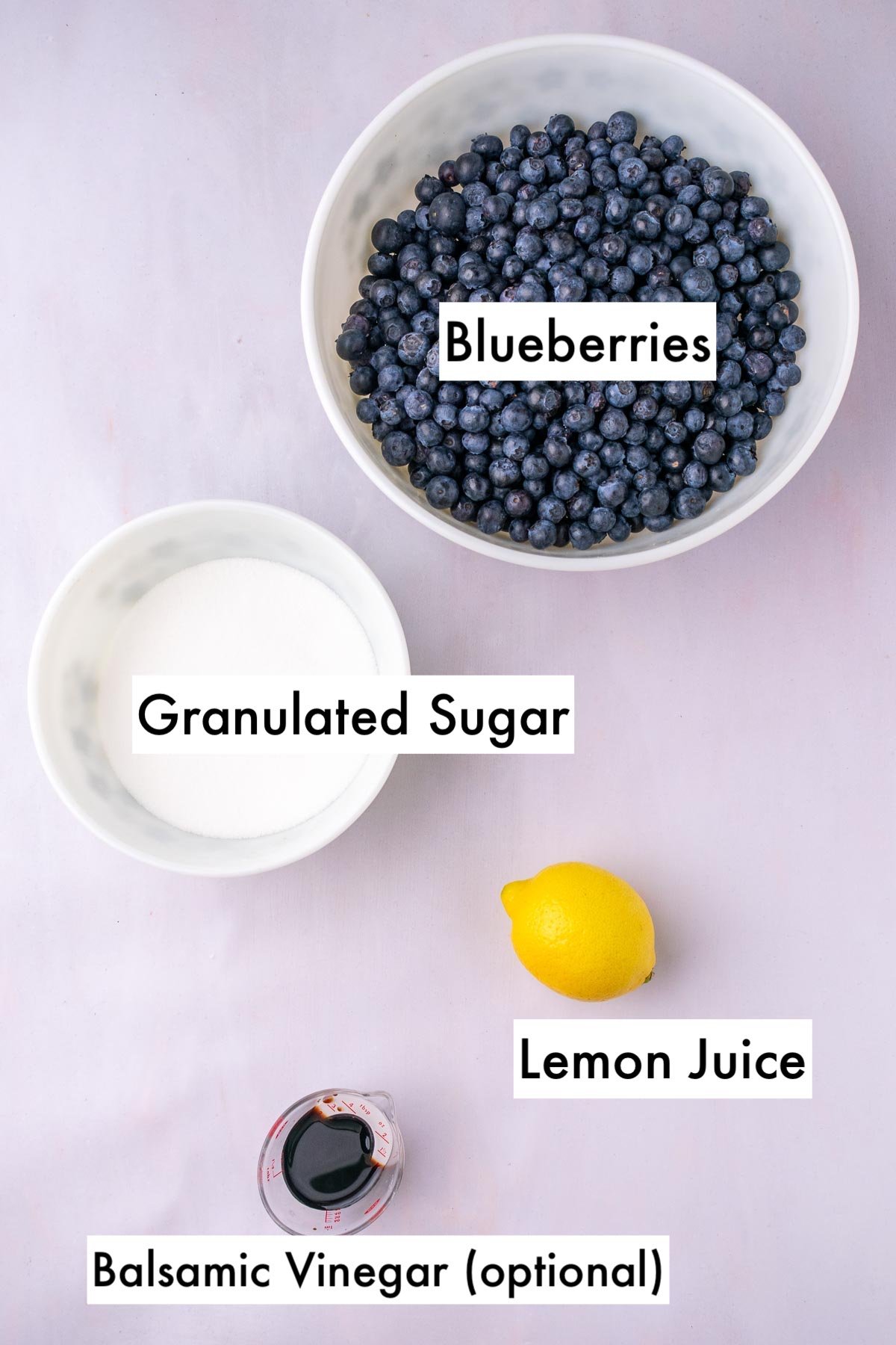 Labeled ingredients needed for blueberry jam including a bowl of blueberries, granulated sugar, lemon juice, and optional balsamic vinegar.