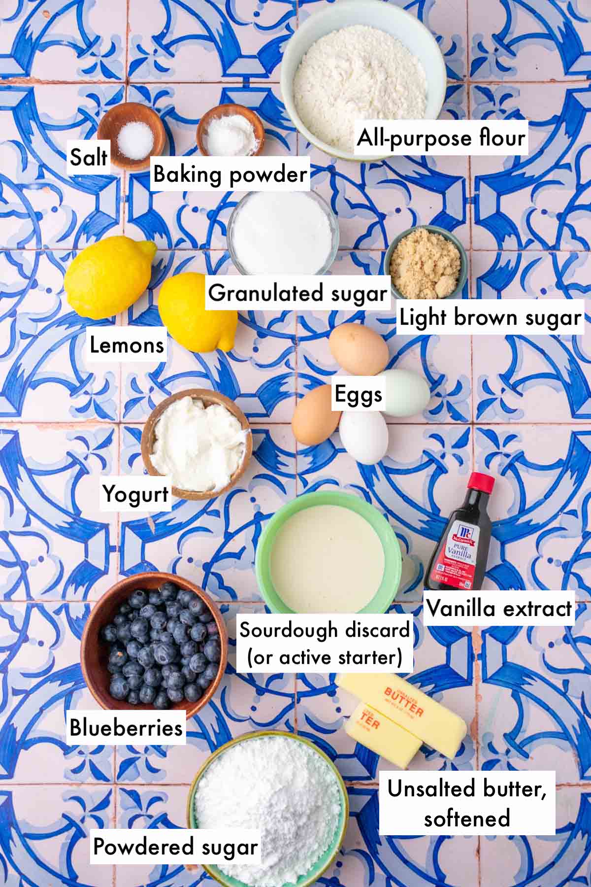 Labeled bowls of ingredients needed for lemon blueberry sourdough pound cake including: all-purpose flour, baking powder, salt, sugar, light brown sugar, lemons, eggs, vanilla, yogurt, sourdough discard, blueberries, powdered sugar, and unsalted butter.