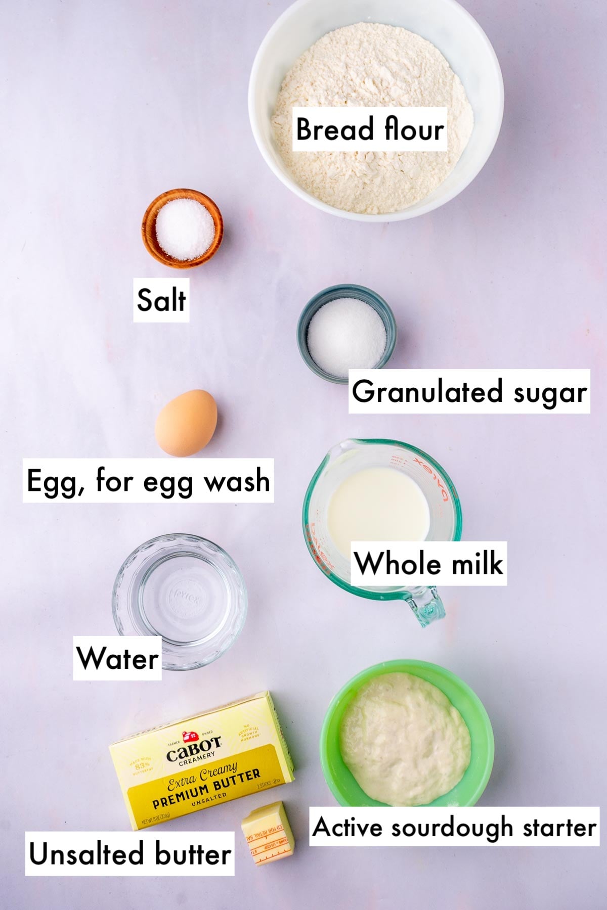 Labeled ingredients needed for sourdough croissant bread loaf including bread flour, salt, sugar, egg for egg wash, whole milk, water, unsalted butter, and active sourdough starter.