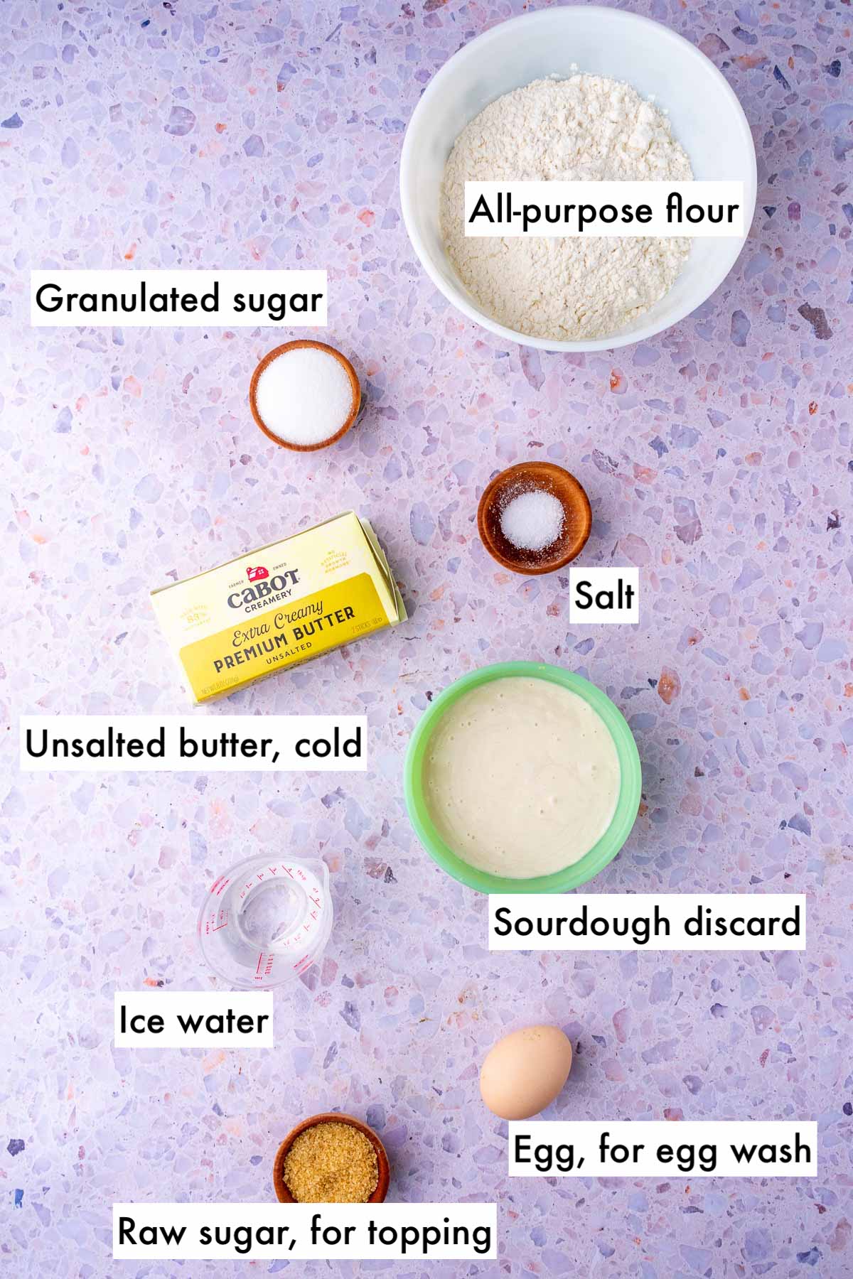 Labeled ingredients needed for sourdough pie crust including all-purpose flour, sugar, salt, unsalted butter, sourdough discard, ice water, egg for egg wash, and raw sugar for topping.