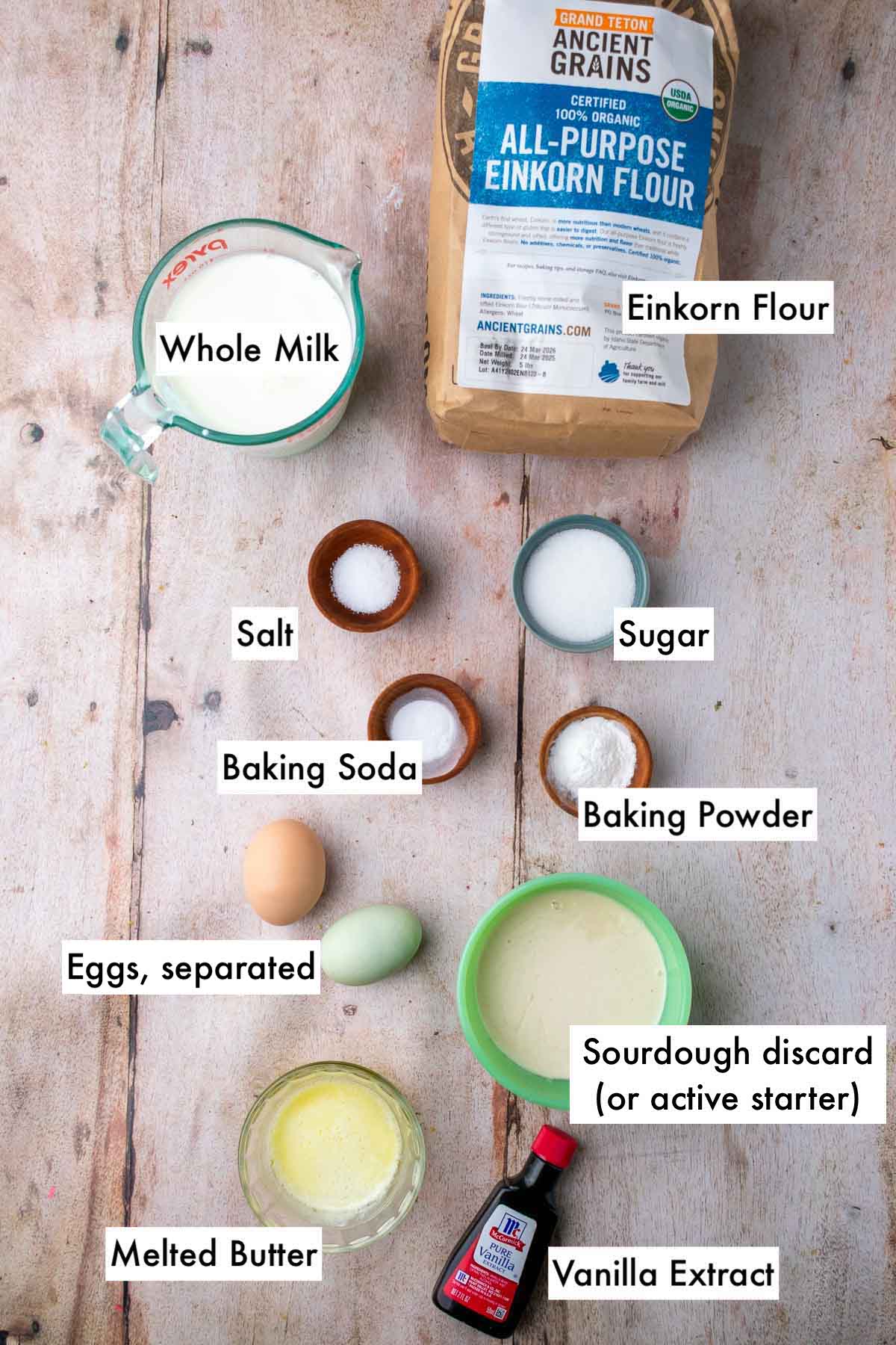 Labeled ingredients needed for sourdough einkorn waffles including einkorn flour, whole milk, sugar, salt, baking powder and soda, eggs separated, sourdough discard, melted butter, and vanilla.