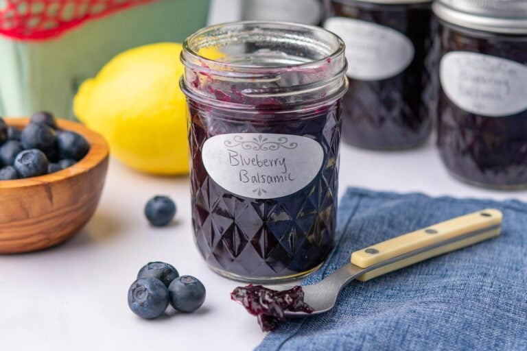 Mason jar with blueberry jam and a spoon with a scoop of jam and more jars with blueberries and a lemon in the background.