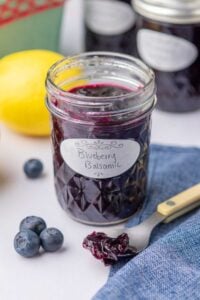 Mason jar with blueberry jam and a scoop of jam on a spoon with blueberries and a lemon in the background.