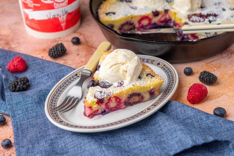 Slice of mixed berry sourdough clafoutis on a plate with ice cream and a fork, surrounded by a cast-iron skillet with clafoutis, a tea towel, berries, and quart of ice cream.