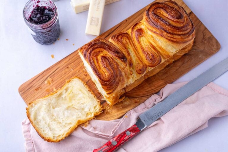 Sourdough croissant bread loaf sliced on a cutting board with a bread knife, jam, and butter.