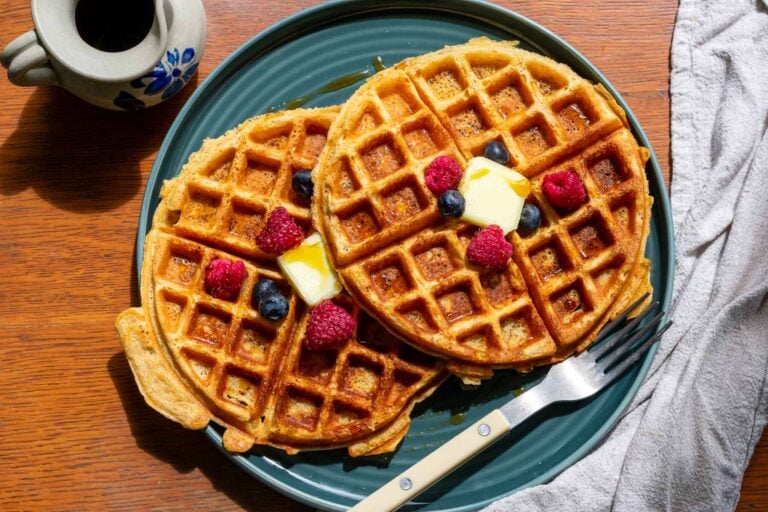 Two sourdough einkorn waffles with butter, maple syrup, and berries on a plate with a fork.