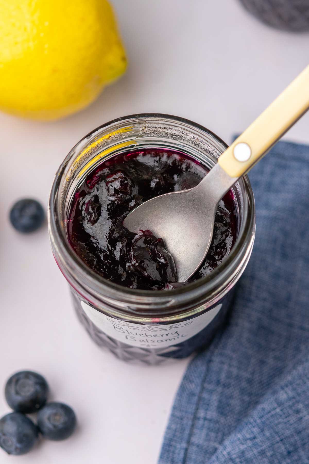 Spoon in a jar with blueberry jam. There's a lemon, blueberries, and a blue towel in the background.