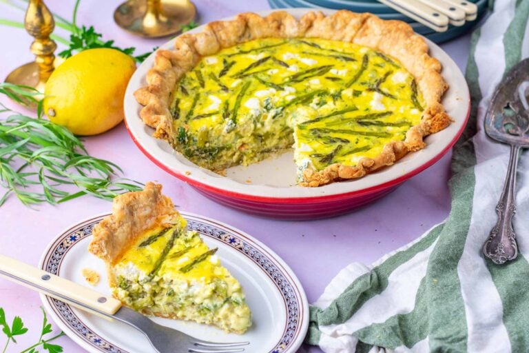 Asparagus goat cheese sourdough quiche in a ceramic pie pan with a slice of quiche on a plate in front. It's surrounded by a lemon, fresh herbs, a pie slicer, and a towel.