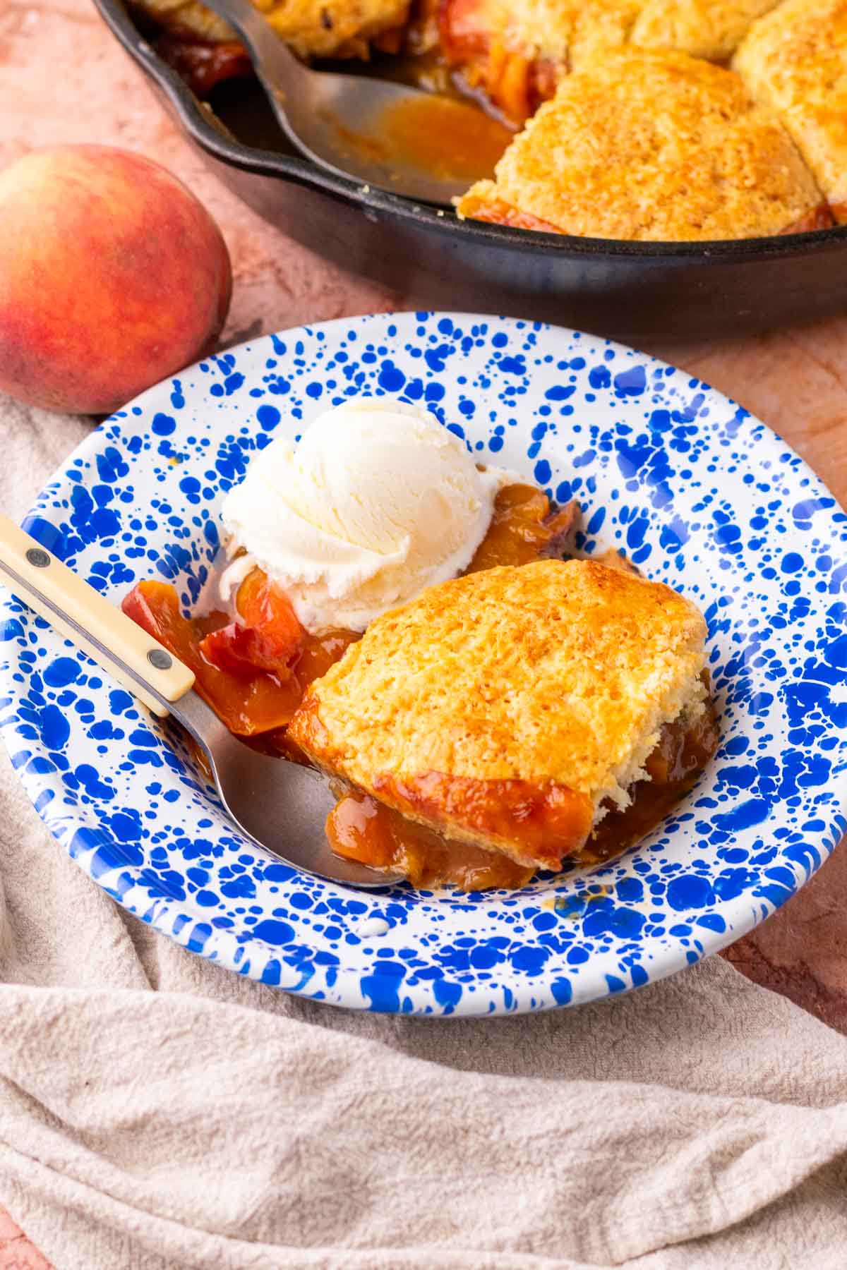 Bowl of sourdough peach cobbler with biscuits, a scoop of ice cream, a spoon, and a peach and skillet in the background.