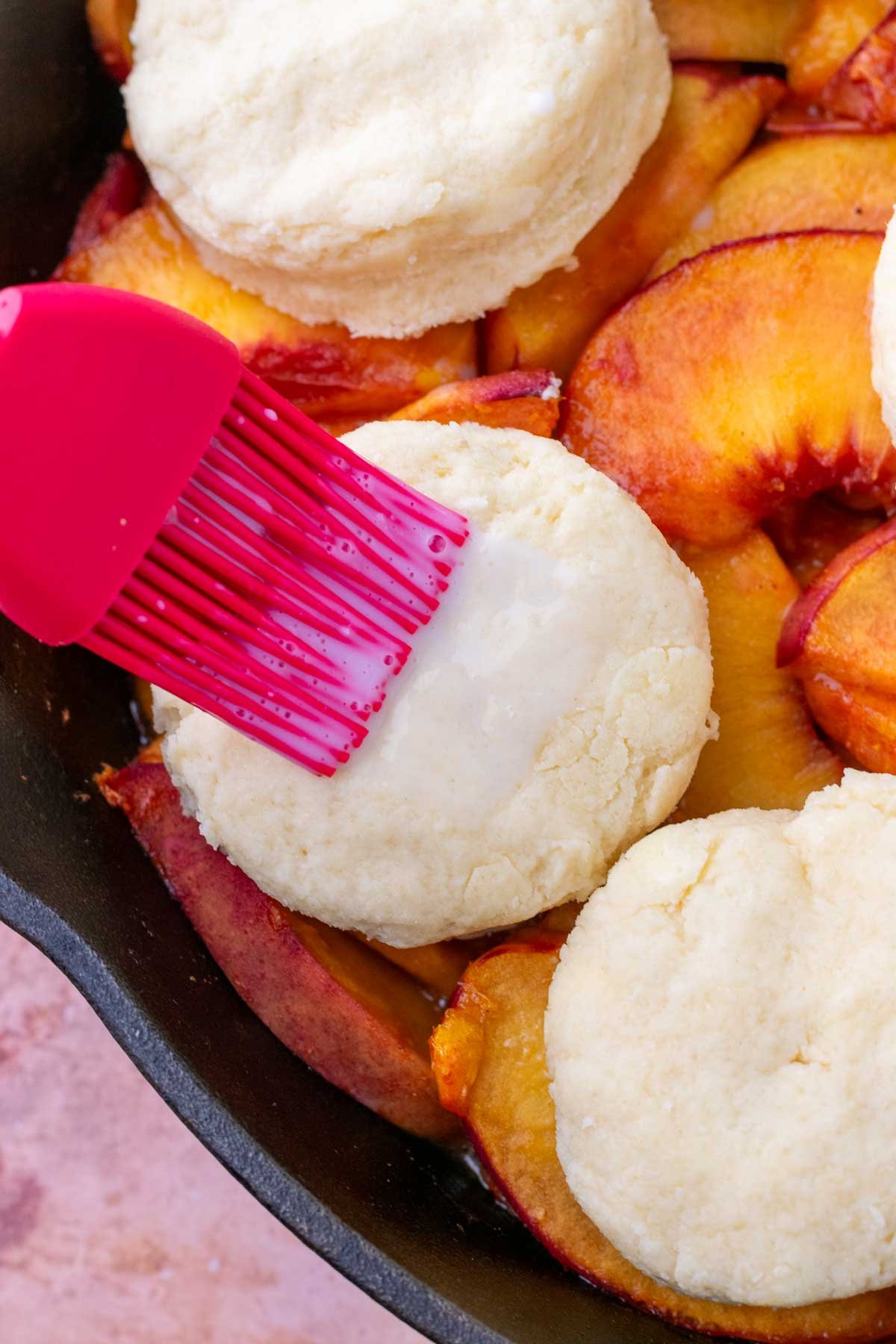 Brushing tops of sourdough biscuits on peach cobbler with buttermilk.
