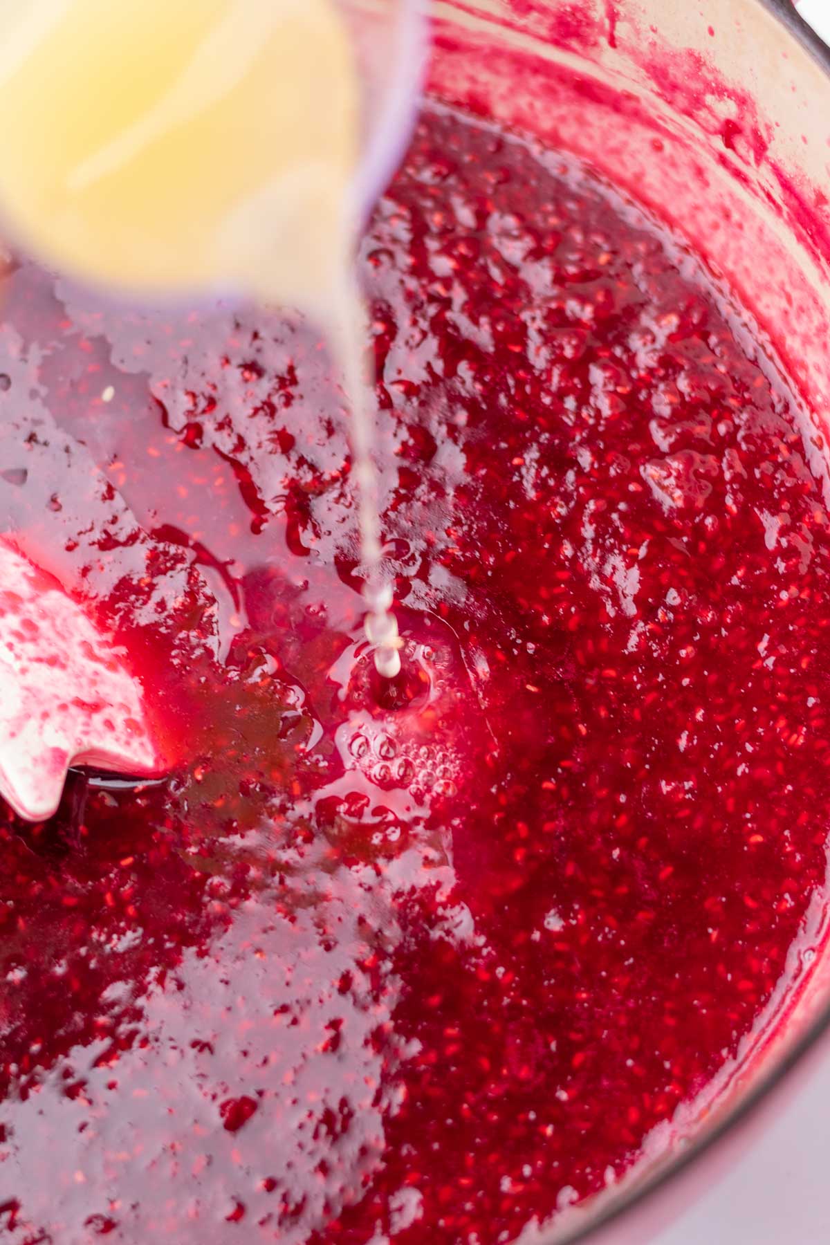 Pouring lemon juice into cooked raspberry jam in a Dutch oven.