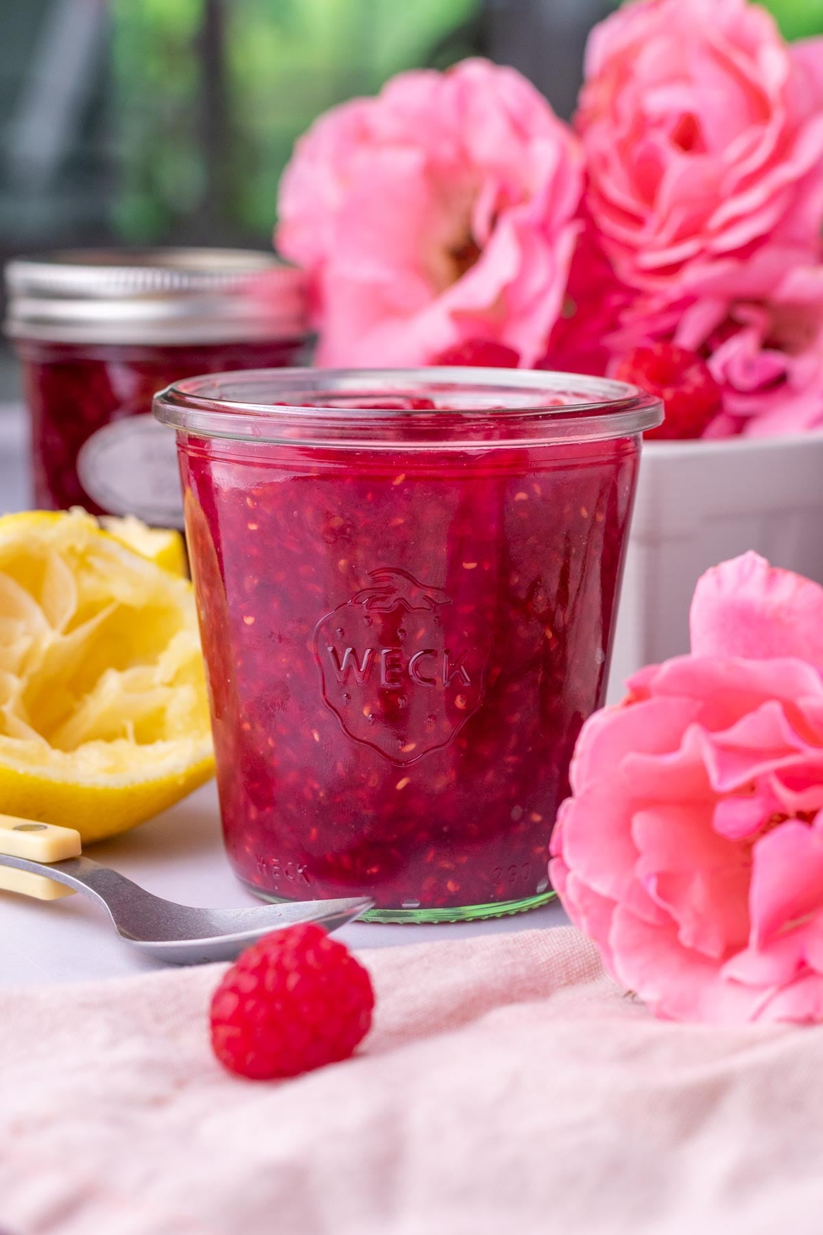 Raspberry Rose Jam in a glass jar surrounded by fresh raspberries and pink roses.