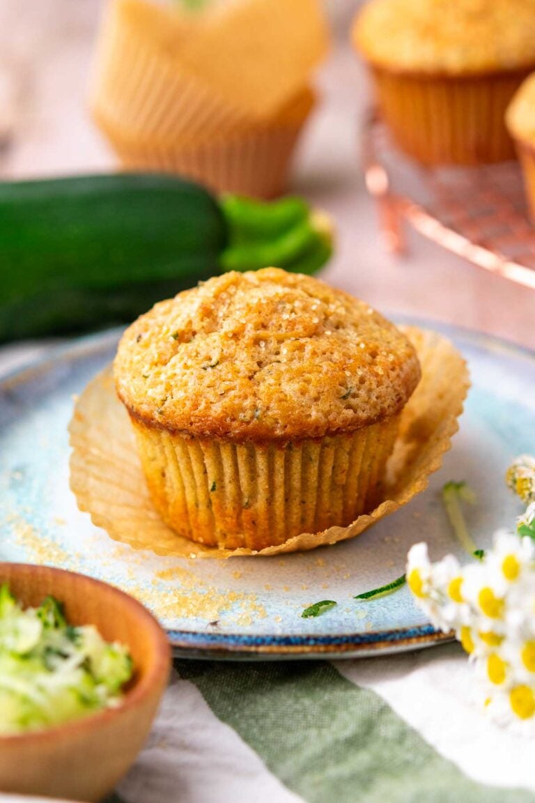 Sourdough zucchini muffin unwrapped from paper liner on a plate with zucchini and more muffins in the background.