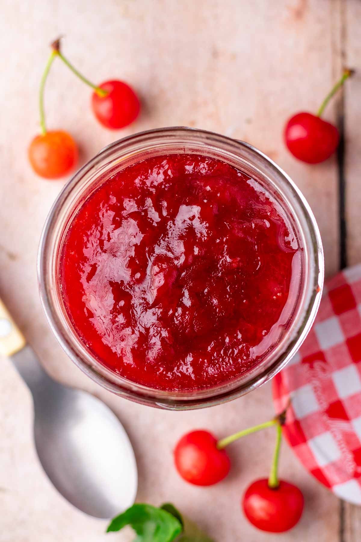 Overhead of the top of a jar of sour cherry jam with a spoon and cherries around it.