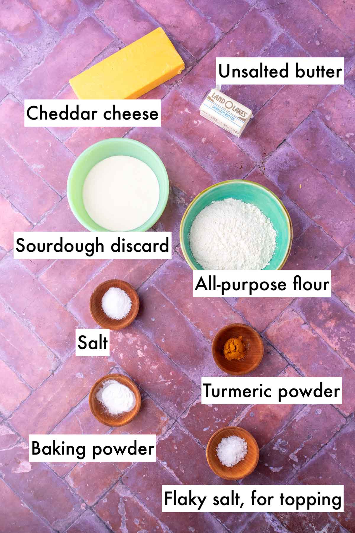 Labeled bowls of ingredients needed for sourdough discard cheese crackers including cheddar cheese, salt, baking powder, sourdough discard, all-purpose flour, unsalted butter, turmeric powder, and flaky salt for topping.
