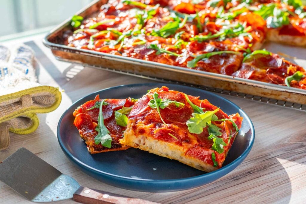 Sourdough pizza al taglio in a sheet pan with two slices on a plate and pepperoni, cheese, and arugula toppings.