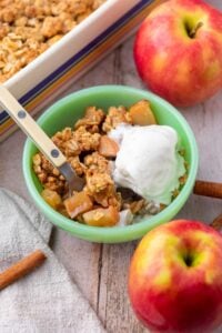 Bowl of sourdough apple crisp with a scoop of ice cream and surrounded by apples.
