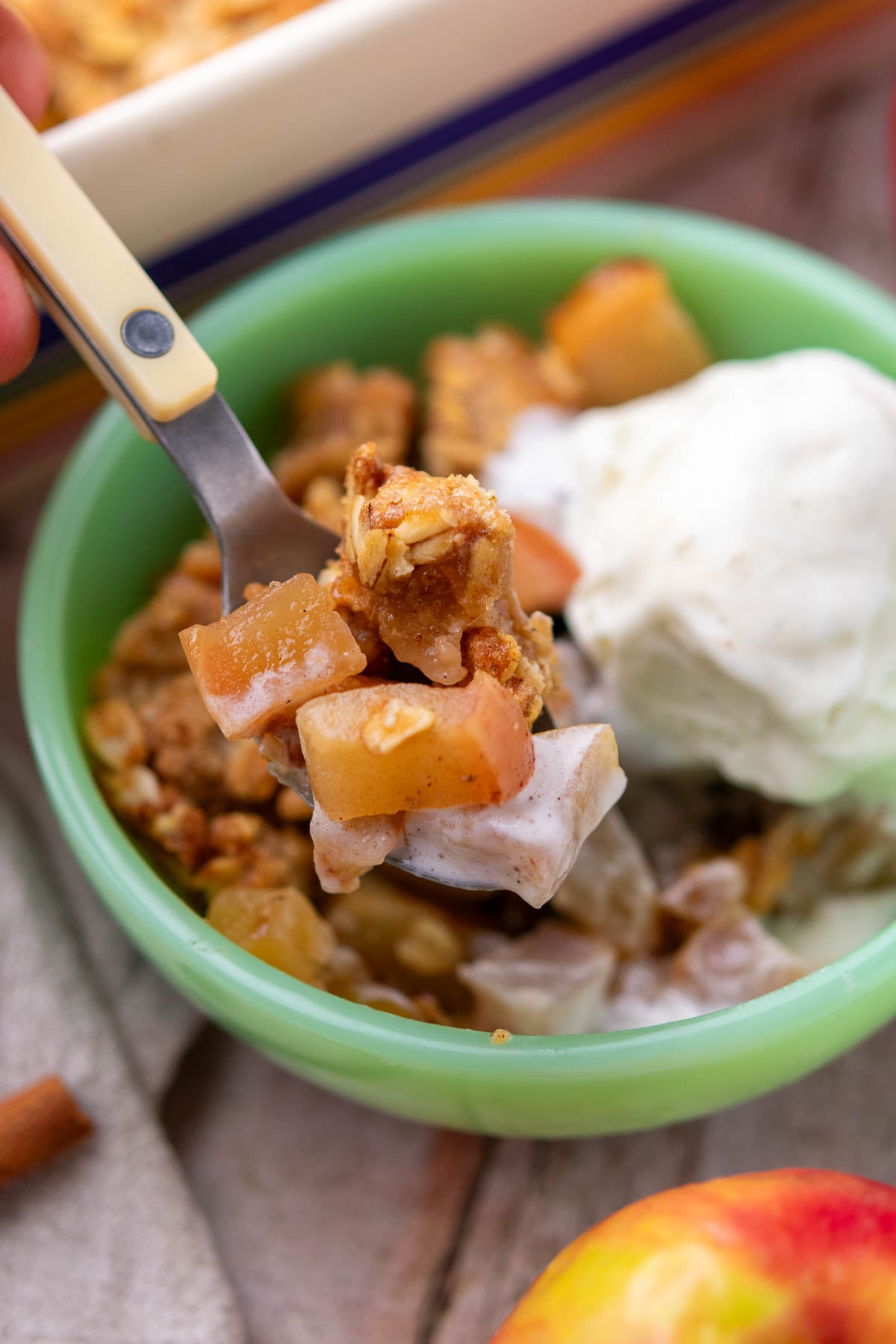 Spoon holding sourdough apple crisp over a bowl with a scoop of ice cream.