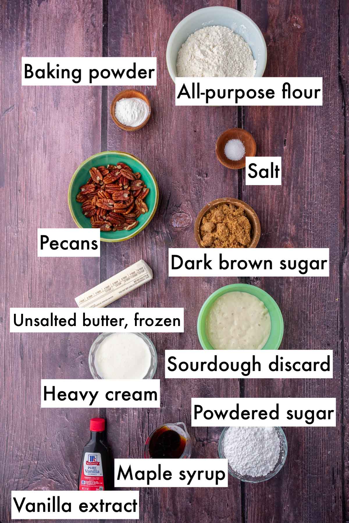 Labeled ingredients needed for sourdough maple pecan scones including all-purpose flour, baking powder, salt, pecans, dark brown sugar, frozen butter, sourdough discard, heavy cream, powdered sugar, maple syrup, and vanilla extract.