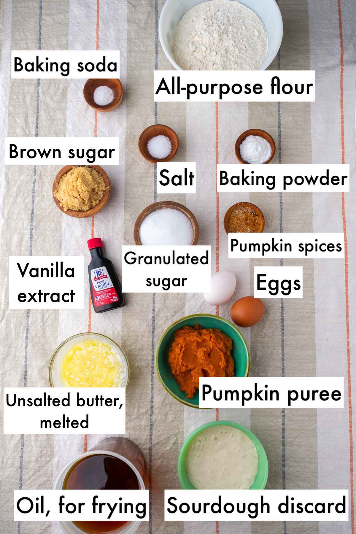 Labeled ingredients needed for sourdough pumpkin donuts, including all-purpose flour, baking powder, baking soda, salt, brown sugar, granulated sugar, eggs, vanilla, pumpkin puree, pumpkin spices, melted butter, frying oil, and sourdough discard.