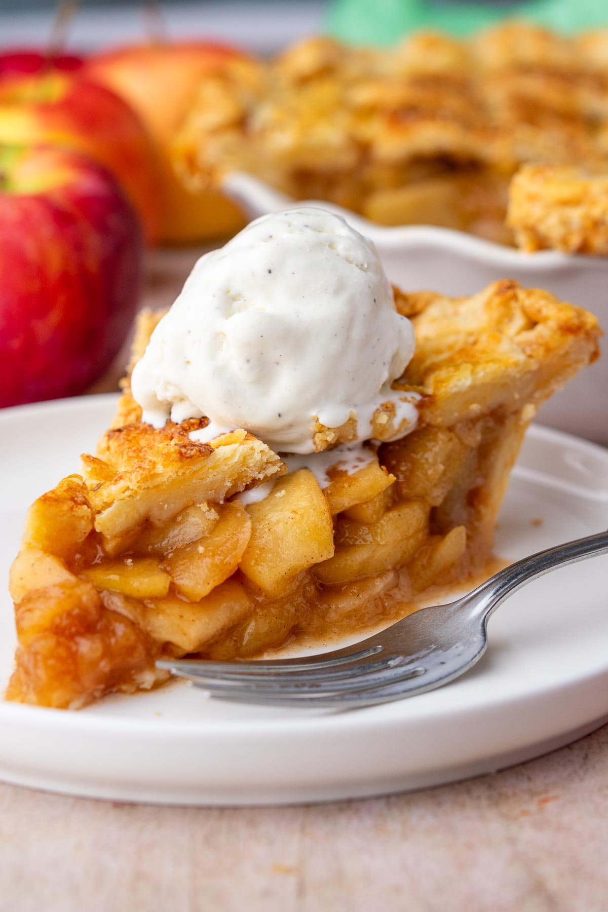 Slice of sourdough apple pie with a scoop of ice cream on a plate with a fork and apples and full pie in the background.