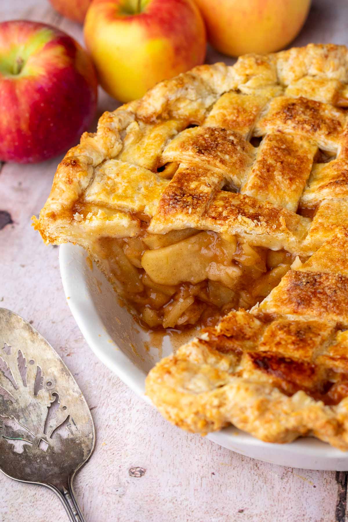 Slice of sourdough apple pie taken out of a pie plate with apples in the background.