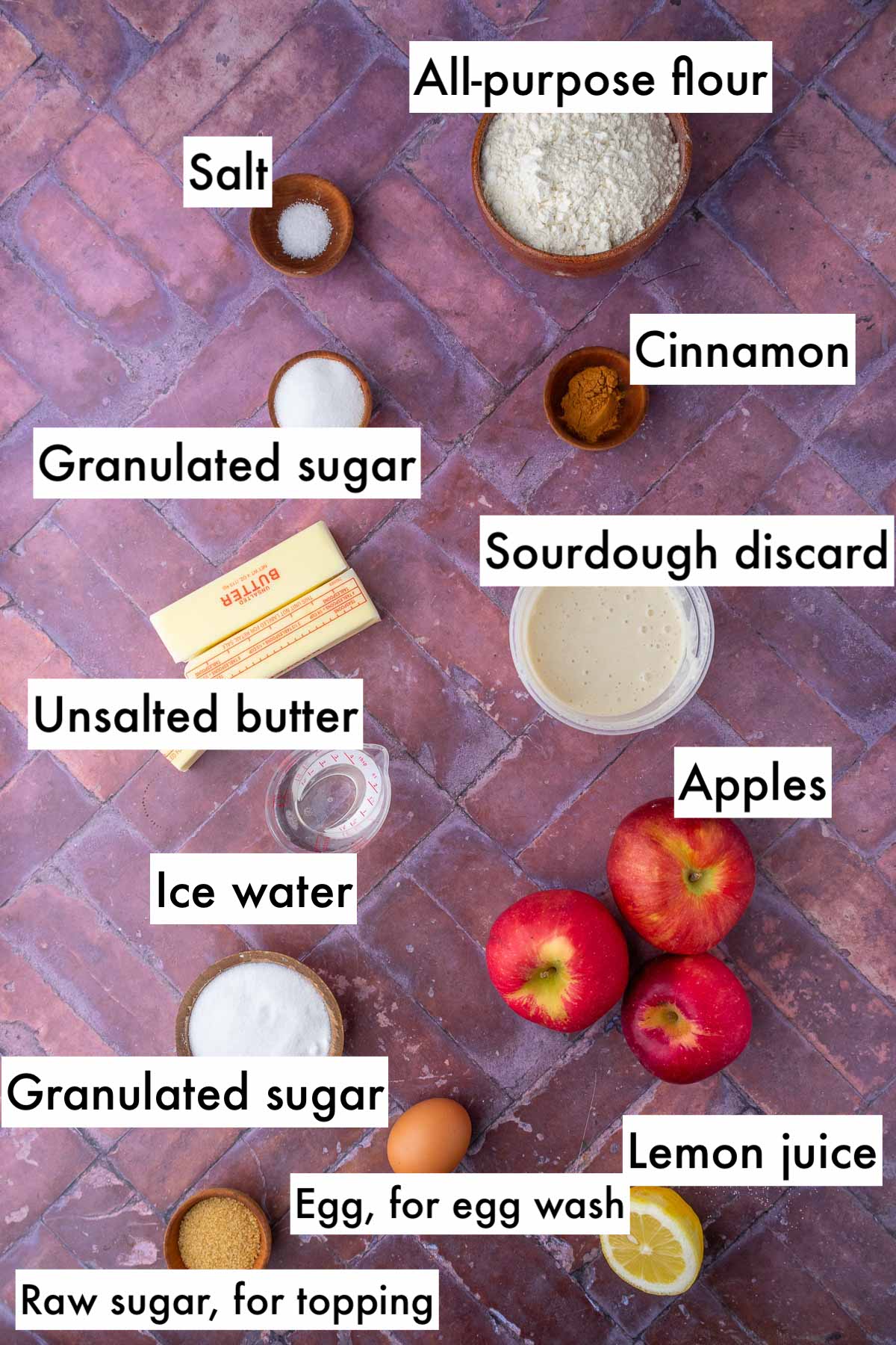 Labeled bowls of ingredients needed for sourdough apple hand pies including all-purpose flour, salt, cinnamon, granulated sugar, sourdough discard, apples, unsalted butter, ice water, egg, lemon juice, and raw sugar.