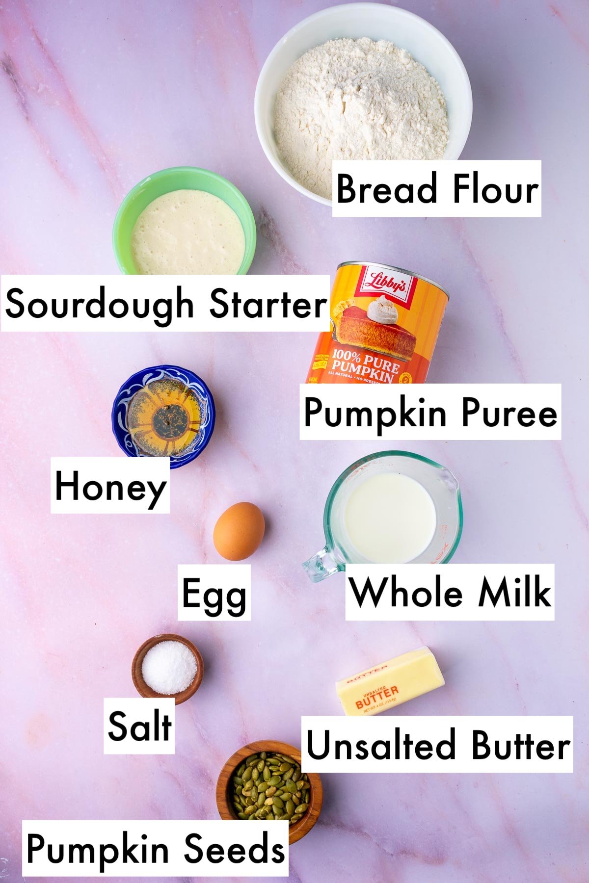 Labeled bowls of ingredients needed for sourdough pumpkin dinner rolls including bread flour, sourdough starter, pumpkin puree, honey, egg, whole milk, salt, unsalted butter, and pumpkin seeds.