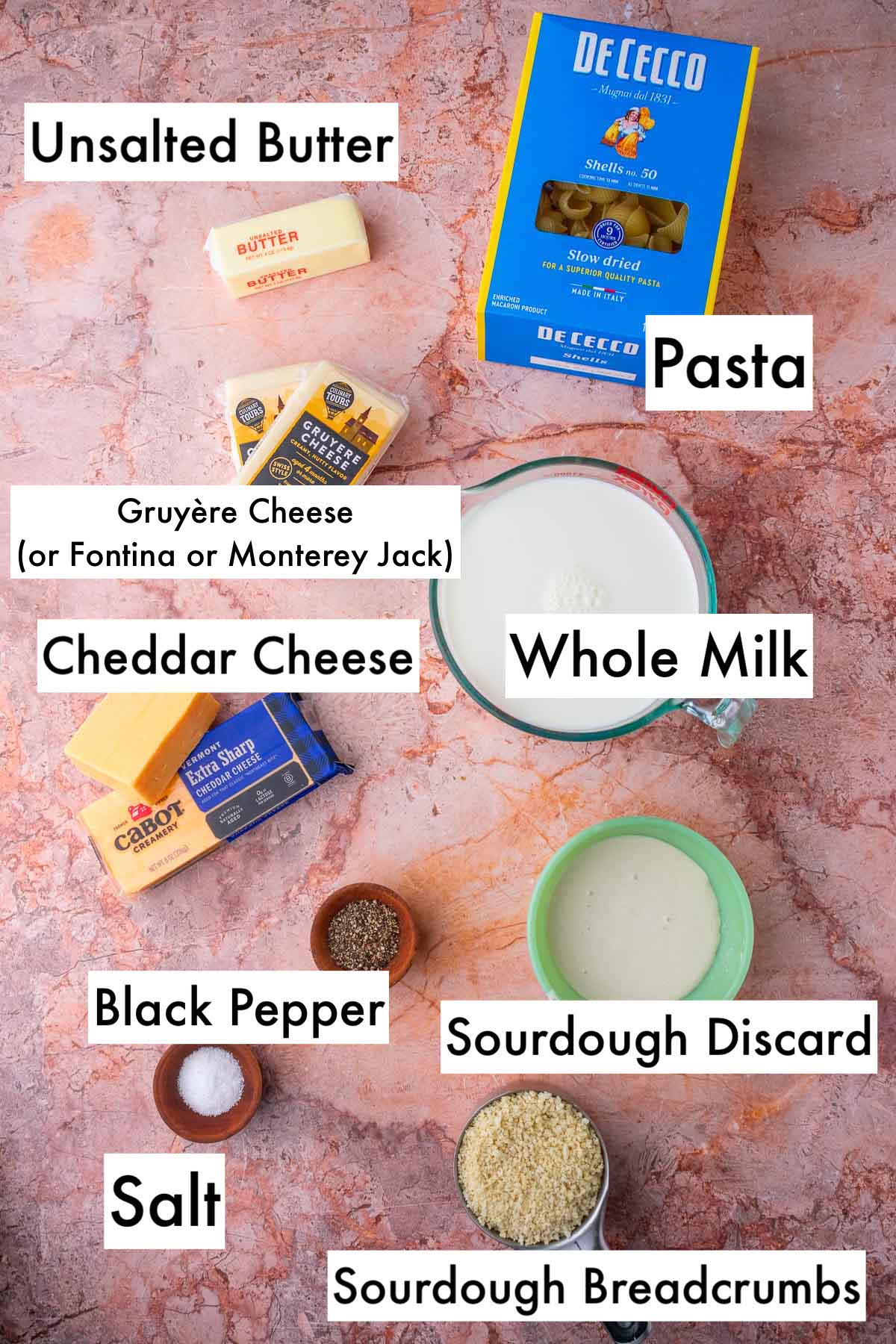 Labeled ingredients needed for sourdough mac and cheese including unsalted butter, pasta, gruyere cheese, cheddar cheese, whole milk, salt and pepper, sourdough discard, and sourdough breadcrumbs.