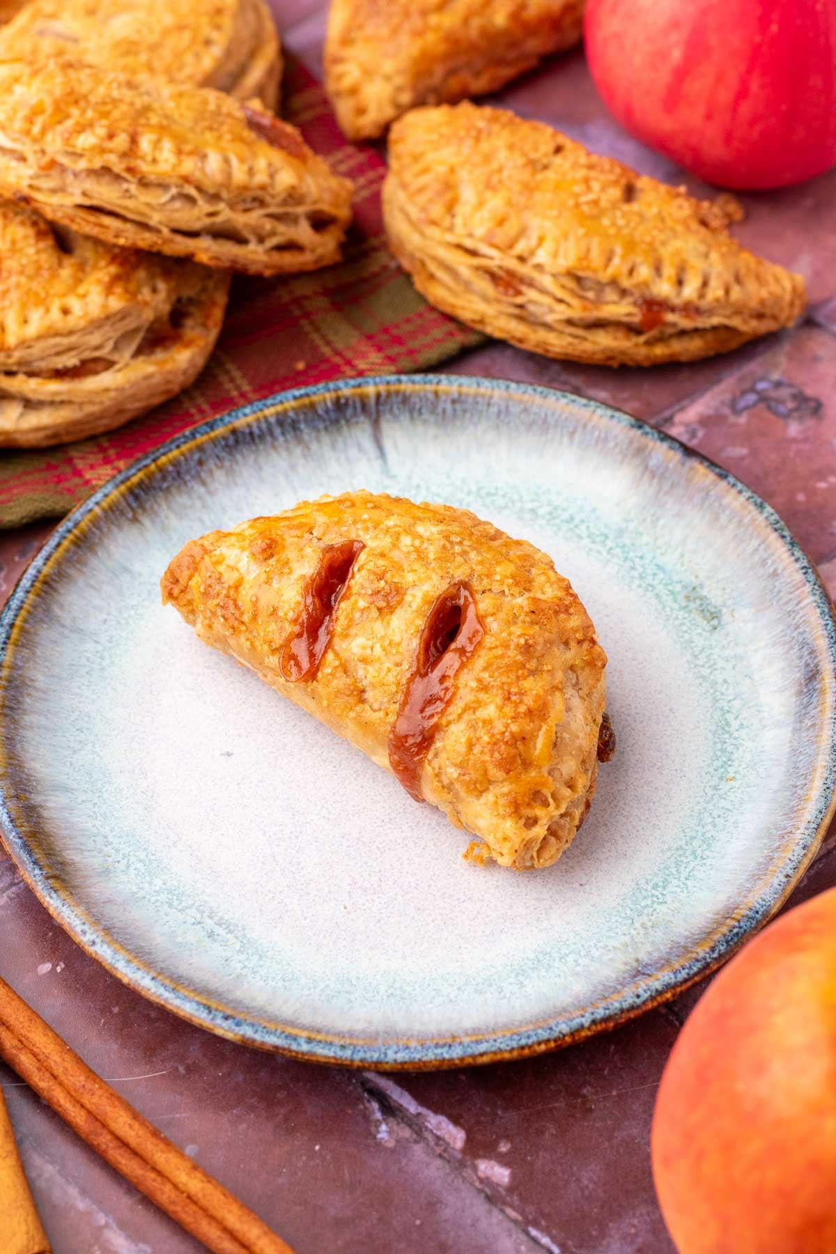 Sourdough apple hand pie on a plate with apples, cinnamon sticks, and more hand pies in the background.