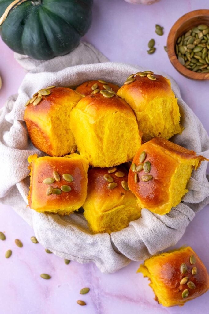 A bowl of sourdough pumpkin dinner rolls with a towel surrounded by a pumpkin and pumpkin seeds.