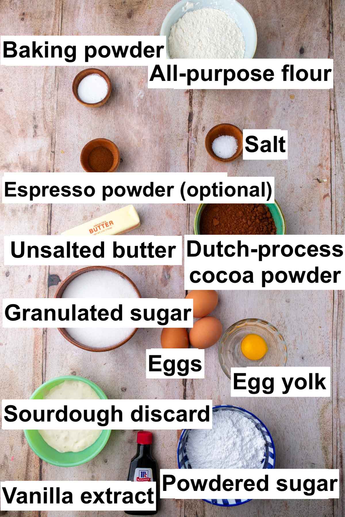 Labeled bowls of ingredients needed for sourdough chocolate crinkle cookies including all-purpose flour, baking powder, salt, cocoa powder, espresso powder, unsalted butter, eggs, egg yolk, granulated sugar, powdered sugar, vanilla extract, and sourdough discard.