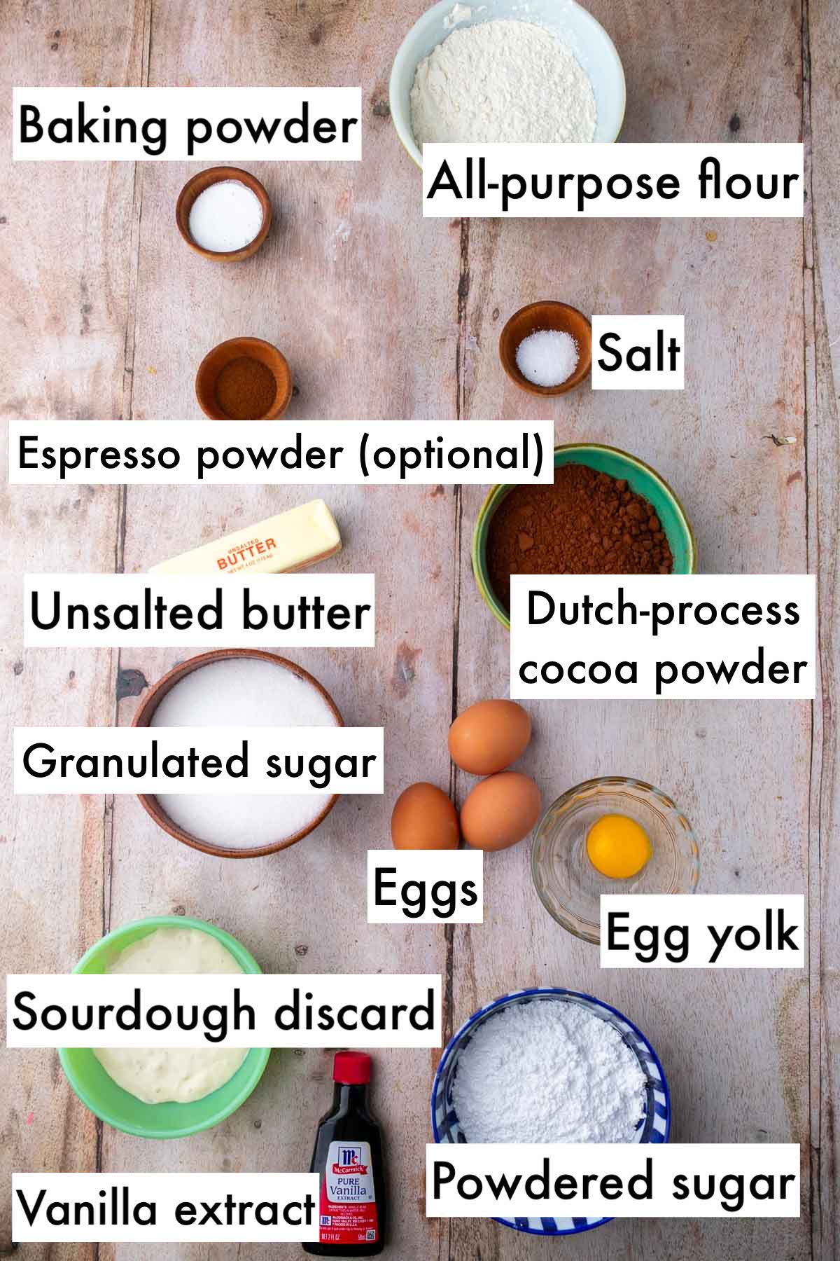 Labeled bowls of ingredients needed for sourdough chocolate crinkle cookies including all-purpose flour, baking powder, salt, cocoa powder, espresso powder, unsalted butter, eggs, egg yolk, granulated sugar, powdered sugar, vanilla extract, and sourdough discard.