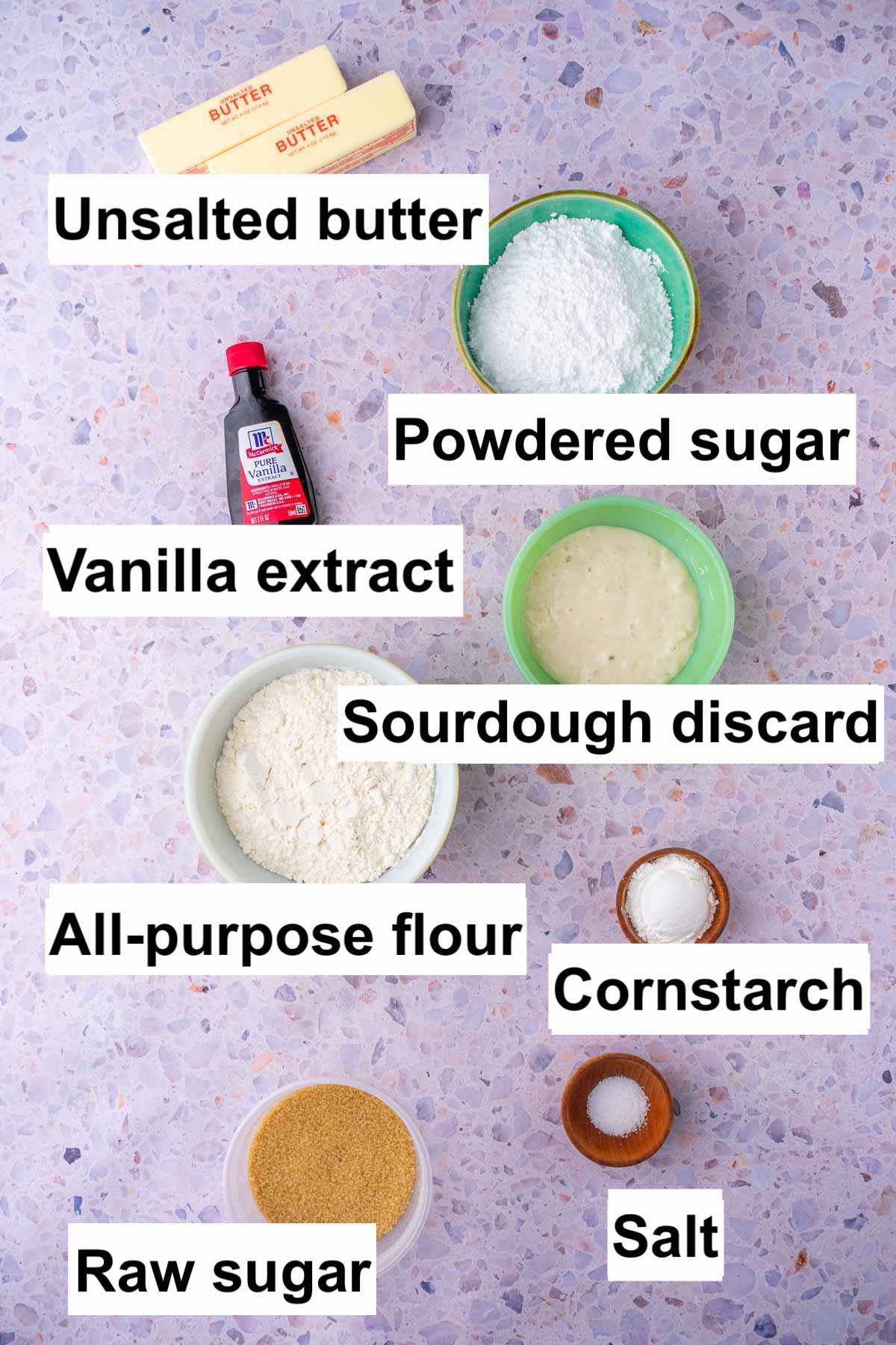 Labeled bowls of ingredients needed for sourdough shortbread cookies including unsalted butter, powdered sugar, vanilla extract, sourdough discard, all-purpose flour, cornstarch, salt, and raw sugar.