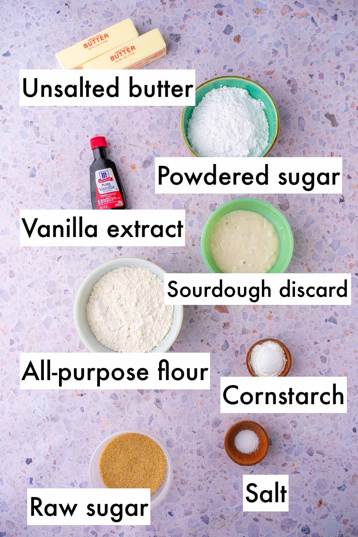 Labeled bowls of ingredients needed for sourdough shortbread cookies including unsalted butter, powdered sugar, vanilla extract, sourdough discard, all-purpose flour, cornstarch, salt, and raw sugar.