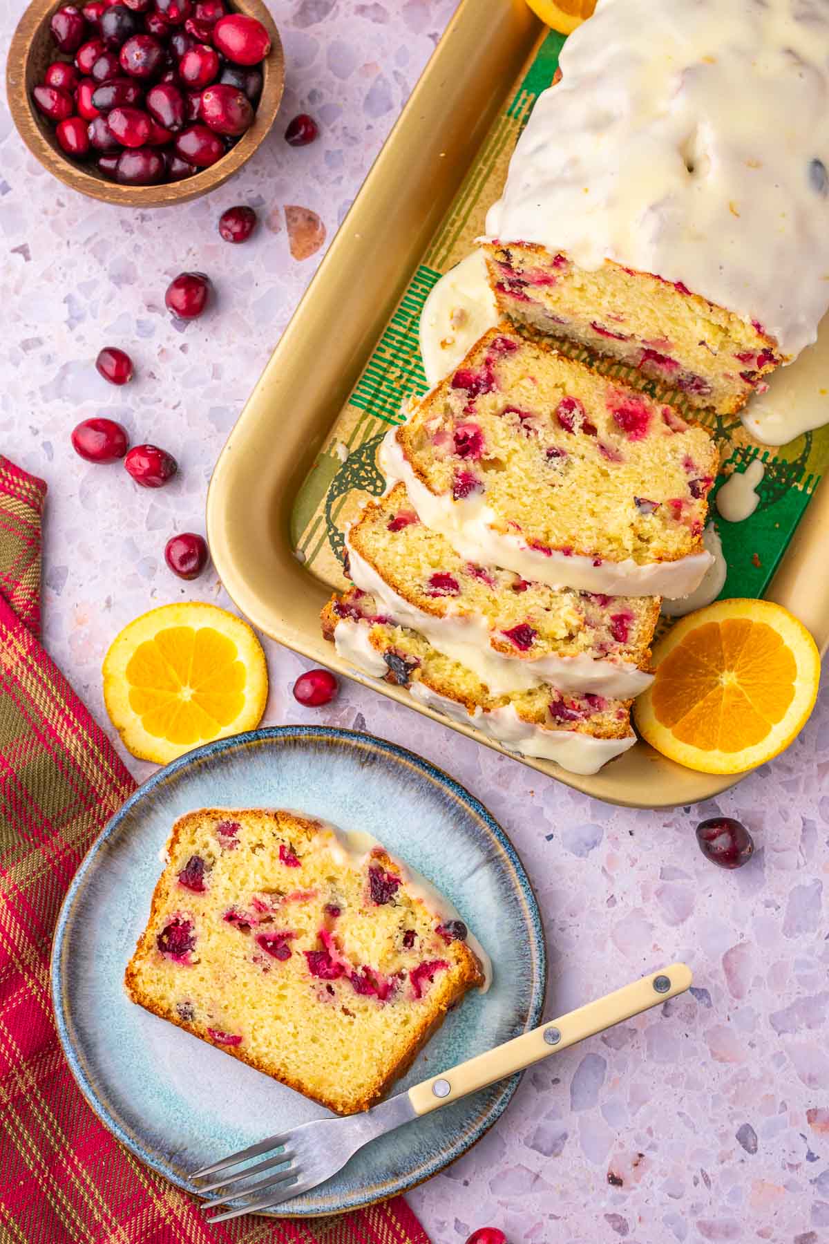 Overhead view of a sliced loaf of Cranberry Orange Sourdough Bread on a platter with a slice on a plate with a fork and surrounded by cranberries and orange slices.