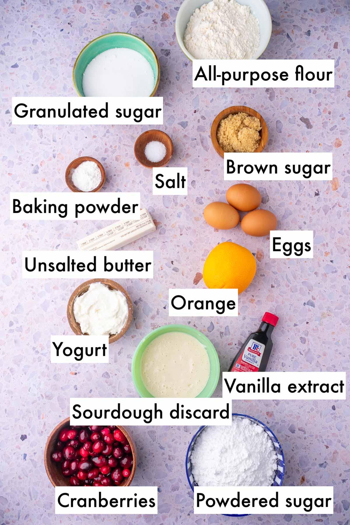 Labeled bowls of ingredients needed for Cranberry Orange Sourdough Bread including all-purpose flour, granulated sugar, brown sugar, salt, baking powder, orange, eggs, unsalted butter, yogurt, vanilla, sourdough discard, cranberries, and powdered sugar.