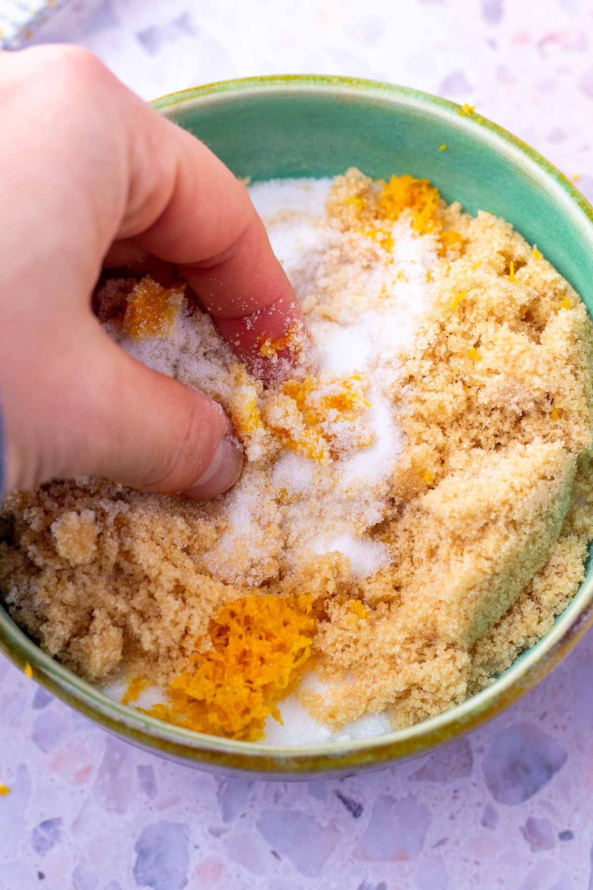Fingers rubbing orange zest into bowl of sugar.