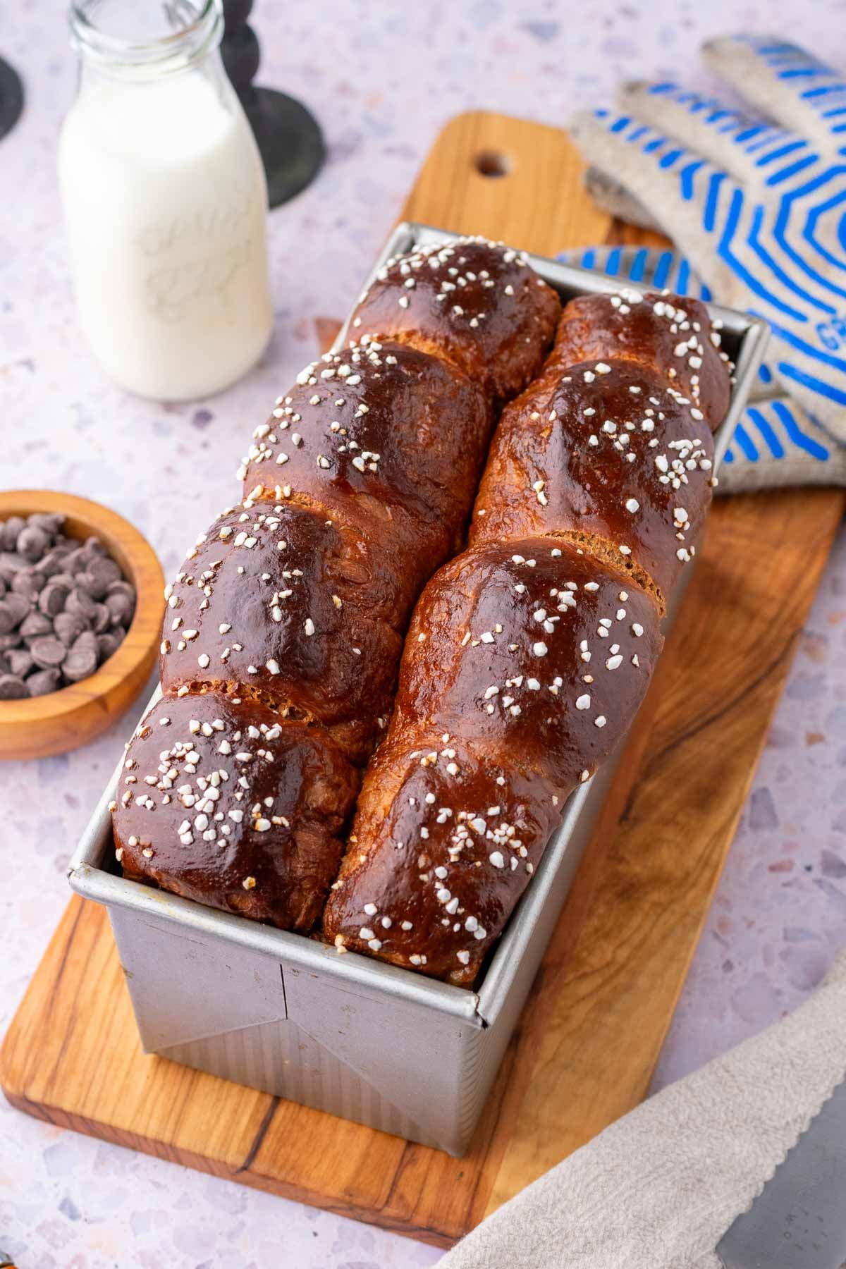 Baked sourdough chocolate brioche bread in a bread loaf pan on a cutting board with gloves, a glass of milk, and a bowl of chocolate chips around it.