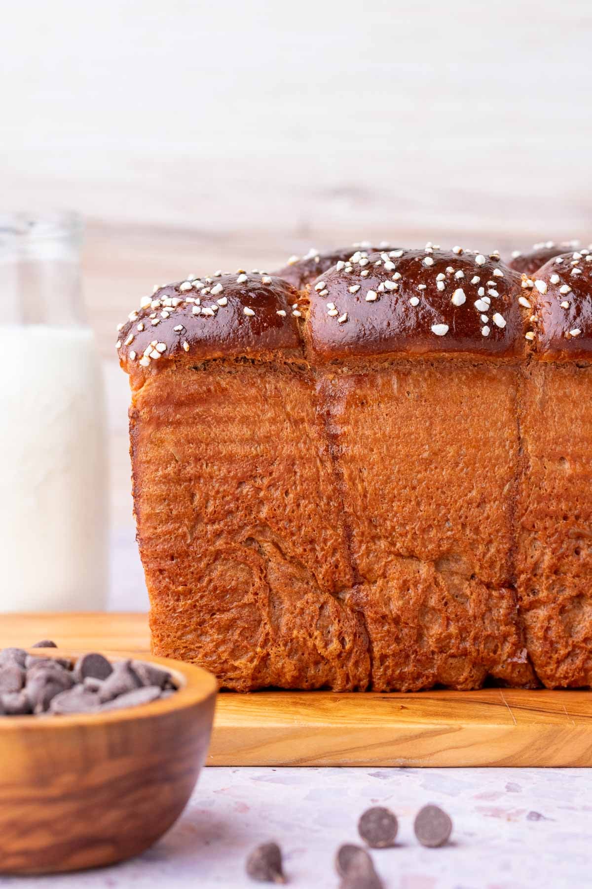 Side profile of fluffy sourdough chocolate brioche bread with a glass of milk in the background and bowl of chocolate chips in front.