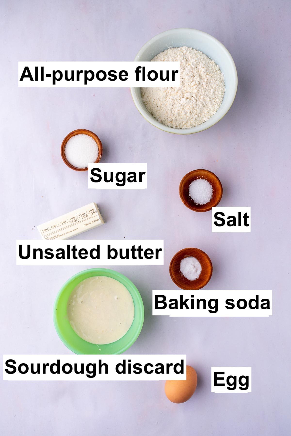 Labeled bowls of ingredients needed for Sourdough Butter Crackers including all-purpose flour, sugar, salt, unsalted butter, baking soda, sourdough discard, and an egg.