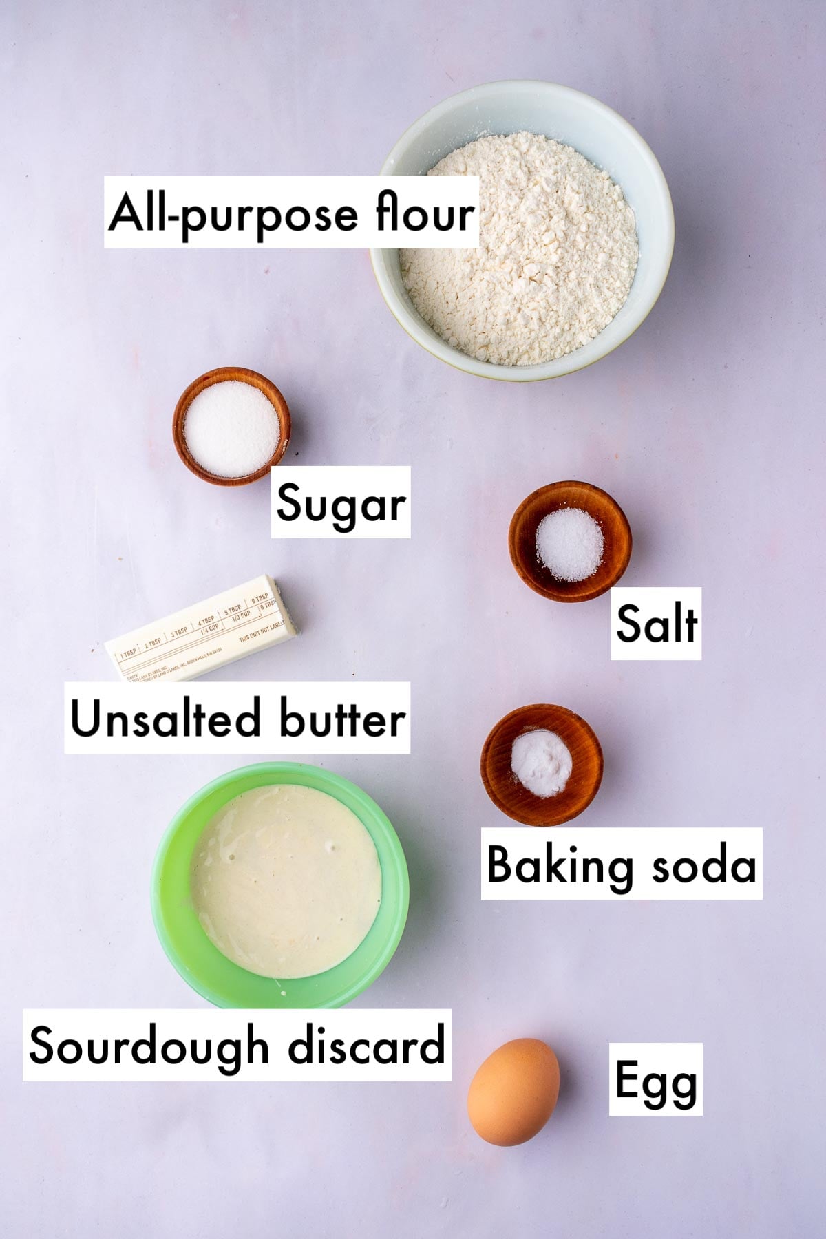 Labeled bowls of ingredients needed for Sourdough Butter Crackers including all-purpose flour, sugar, salt, unsalted butter, baking soda, sourdough discard, and an egg.