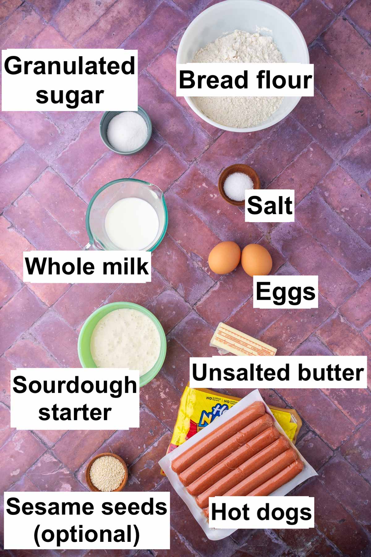 Labeled bowls of ingredients needed for sourdough pigs in a blanket including bread flour, granulated sugar, salt, hot dogs, eggs, whole milk, sourdough starter, unsalted butter, and optional sesame seeds.