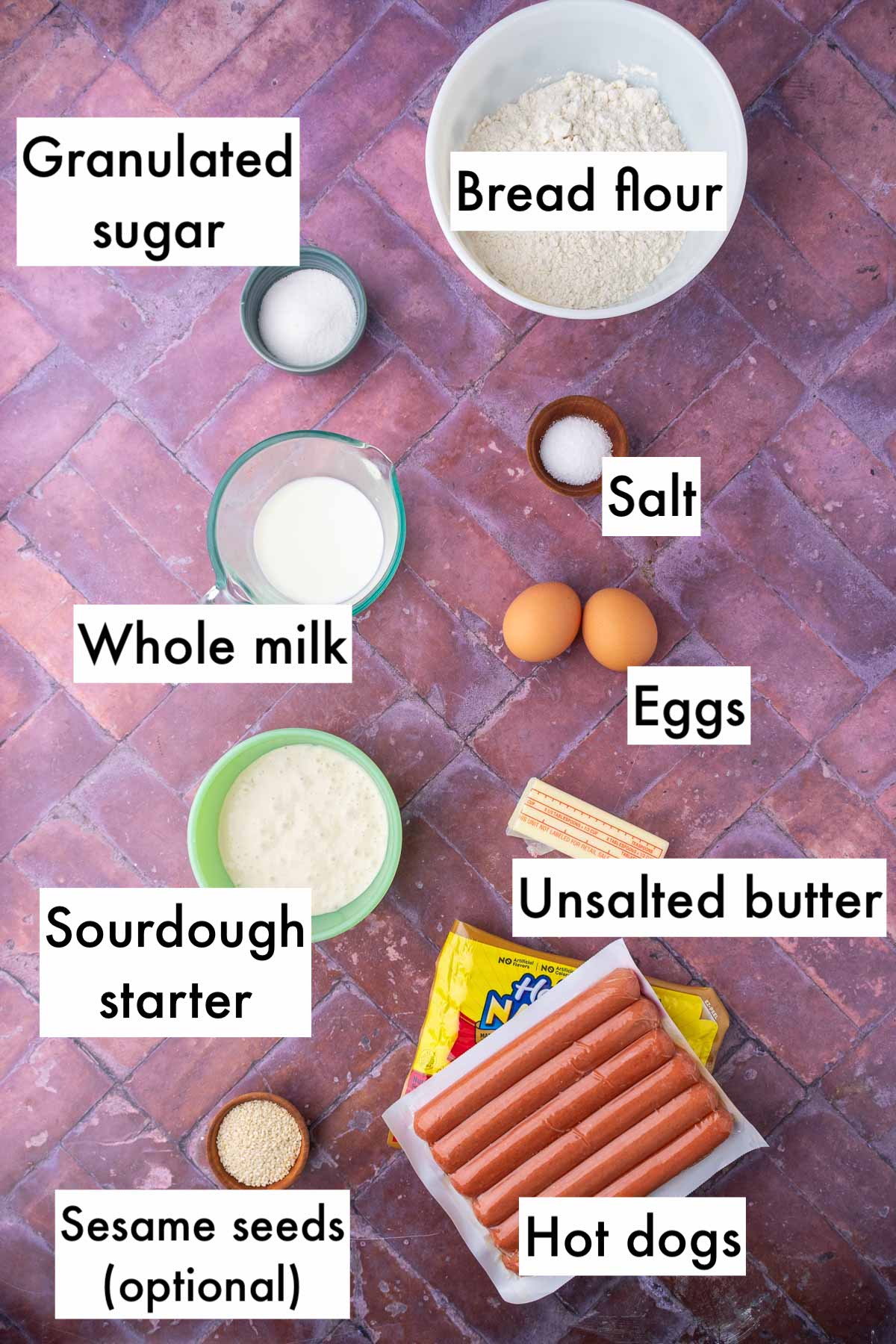 Labeled bowls of ingredients needed for sourdough pigs in a blanket including bread flour, granulated sugar, salt, hot dogs, eggs, whole milk, sourdough starter, unsalted butter, and optional sesame seeds.